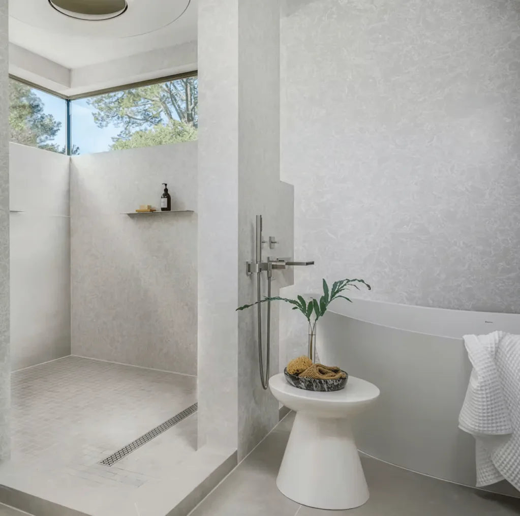 Modern bathroom with a walk-in shower featuring frameless glass and light gray stone tiles, a freestanding white bathtub, and a small white side table holding a plant and natural sponges.