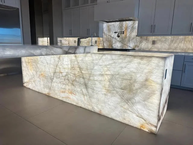 Modern kitchen interior featuring a large illuminated quartz countertop island with subtle veining, white cabinetry, stainless steel appliances, and a minimalist design.