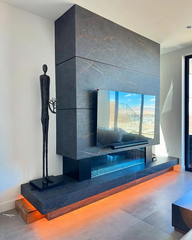 Modern living room with a sleek dark stone fireplace and mantel featuring integrated LED lighting and a mounted flat-screen TV, accompanied by a tall, slender abstract sculpture on one side.