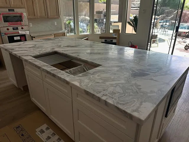 Large kitchen island with polished light gray and white marble quartzite countertop featuring an undermount sink cutout, set against light wood cabinetry and glass doors overlooking an outdoor patio.