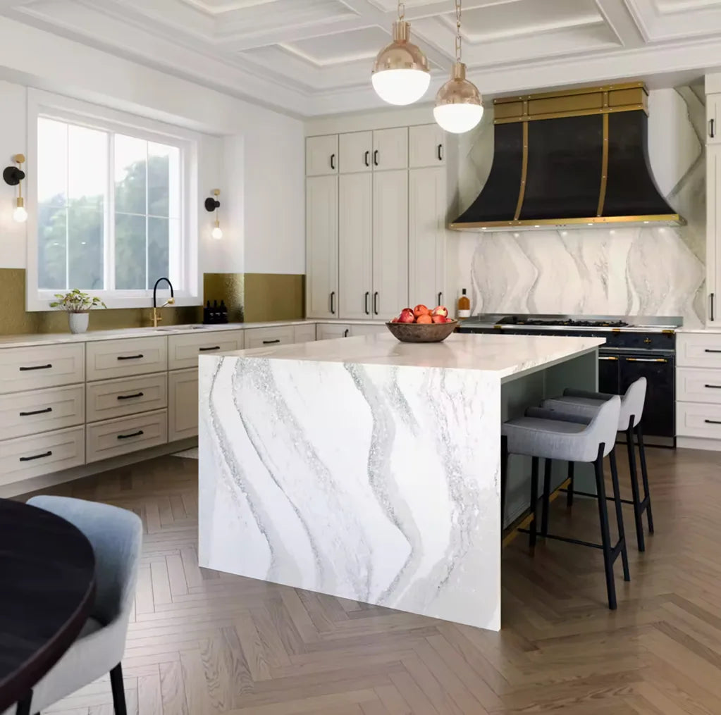 Modern kitchen interior featuring white quartz countertops with gray veining, matching island with seating, light cabinetry, large window, brass-accented range hood, and wood herringbone flooring.