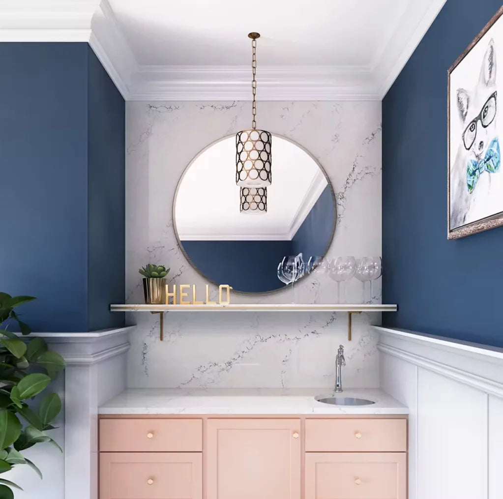 Modern bathroom vanity with soft pink cabinets, a white countertop with a small round sink, blue walls, decorative round mirror, hanging pendant light, and a "HELLO" sign on a floating shelf.