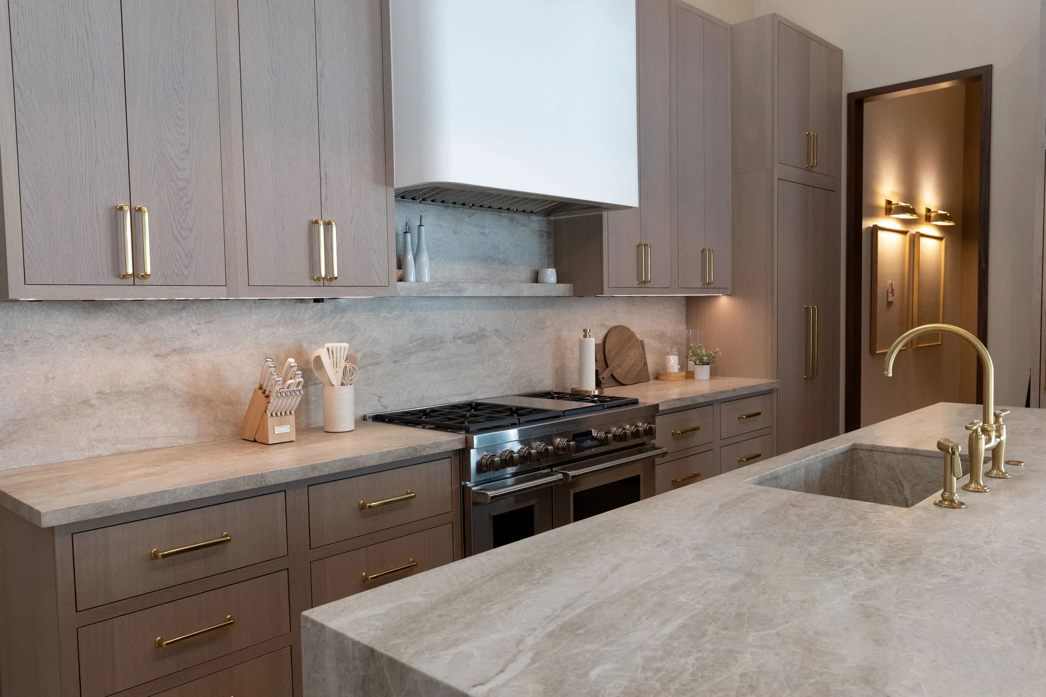 Modern kitchen with light wood cabinets, gold handles, and marble countertops for a polished look. A built-in stove, white range hood, and matching island with gold faucet offer style reminiscent of luxurious quartz countertops. Utensils and decor are neatly arranged.