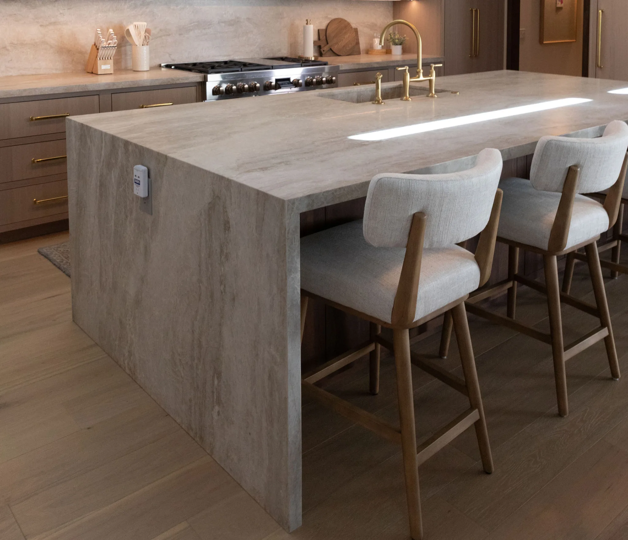 Modern kitchen with a large marble island and sleek marble countertops, gold faucet, and two cushioned bar stools. Light wood floors and drawers with gold handles complement the stove and countertop accessories in the background.