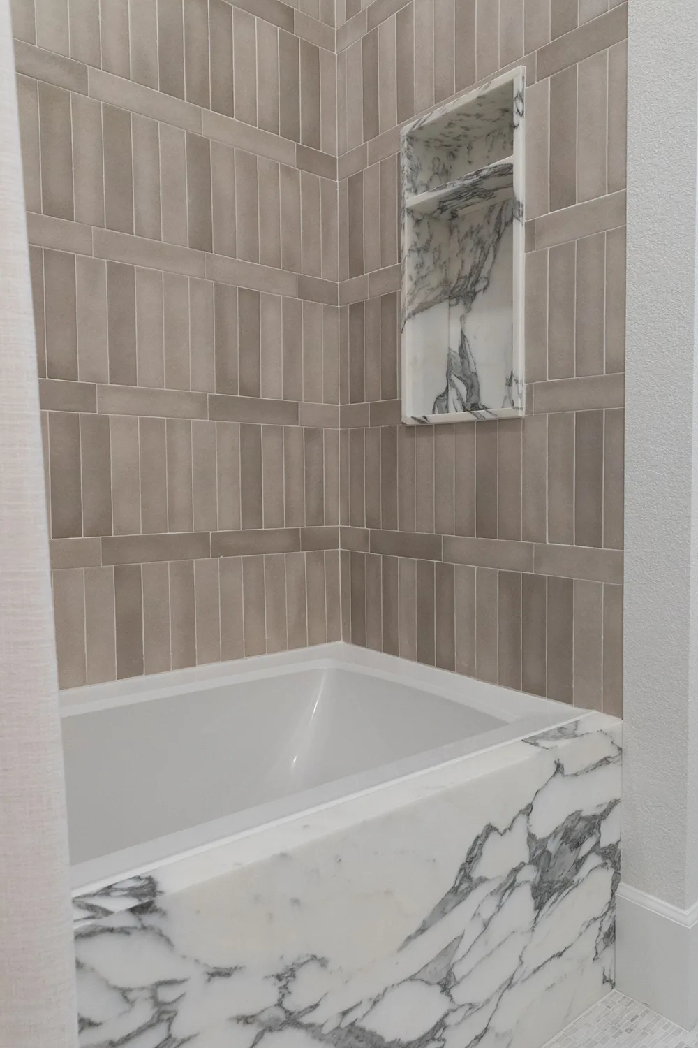A modern bathtub with a white and gray marble front panel, brown rectangular wall tiles, and a built-in niche lined with matching marble above the tub—echoing the elegance often seen in bathroom countertops.