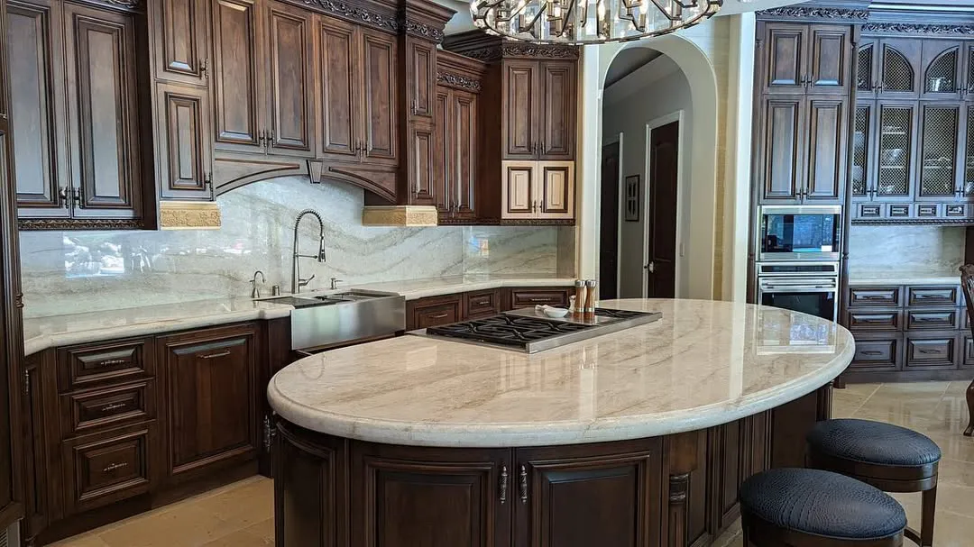 A luxurious kitchen with dark wooden cabinets, marble countertops, a farmhouse sink, a large oval island with a cooktop and two round cushioned stools, featuring elegant lighting and built-in appliances for stunning kitchen countertops.