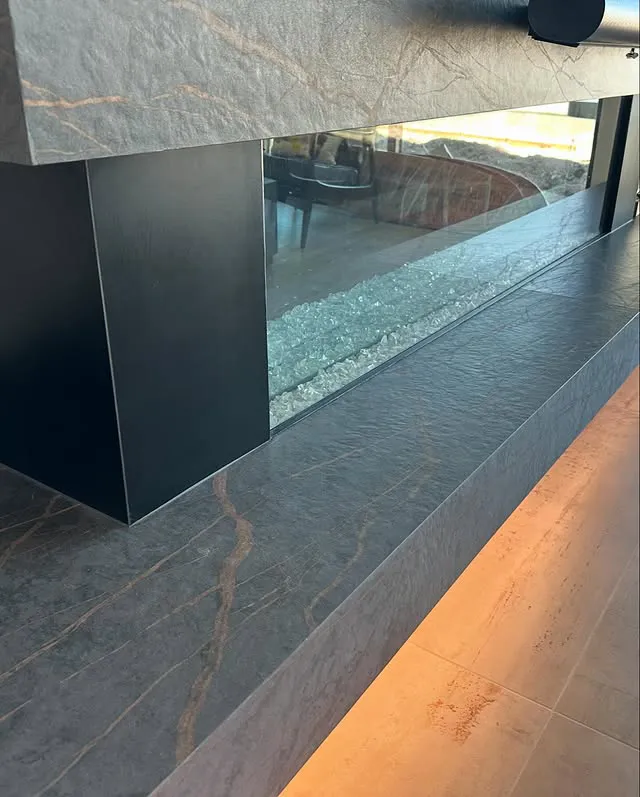 Close-up of a modern glass and stone fireplace with a sleek black metal frame, textured gray stone surfaces reminiscent of premium kitchen countertops, and warm accent lighting along the floor.