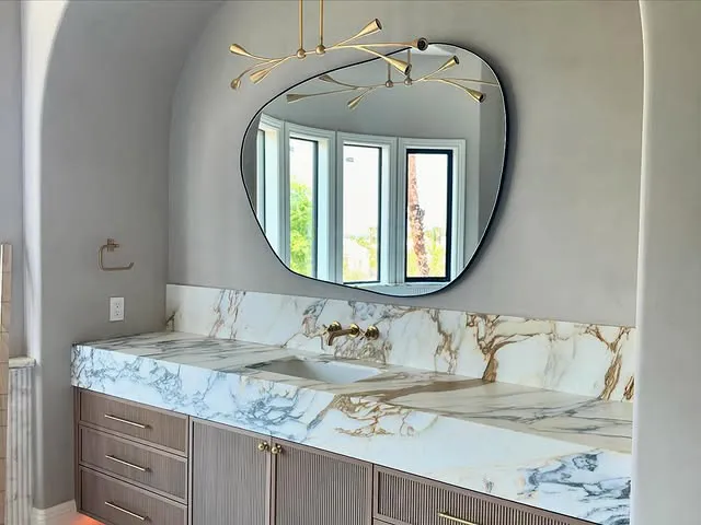 Modern bathroom vanity with a quartz countertop, gold fixtures, a large irregular-shaped mirror, light wood cabinets, and a window reflected in the mirror. A contemporary chandelier hangs overhead—a Signature Stone Las Vegas design.