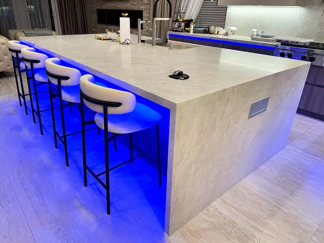 A modern kitchen island with five white bar stools, marble kitchen countertops, and blue LED lighting underneath. The kitchen features a stove, sink, and various items on the counter, all in a neutral color palette by Signature Stone Las Vegas.