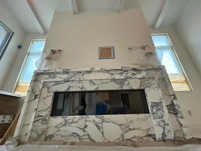 A modern fireplace with a black glass opening and a white and gray marble surround, reminiscent of elegant kitchen countertops, set against a tall, light-colored wall. Large windows on either side let in natural light; the room appears to be under construction.