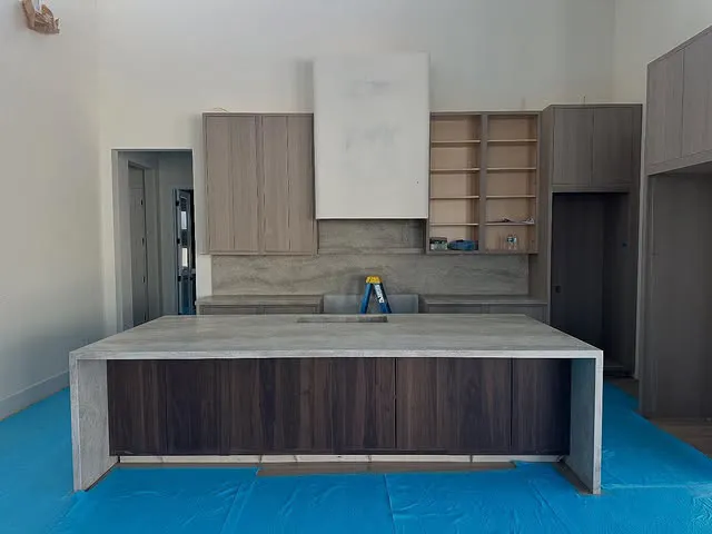 A modern kitchen under construction with a large island, open shelves, and wood cabinets. Blue protective sheets cover the floor, while tools and materials rest on the countertop, ready for the installation of stunning quartz countertops.