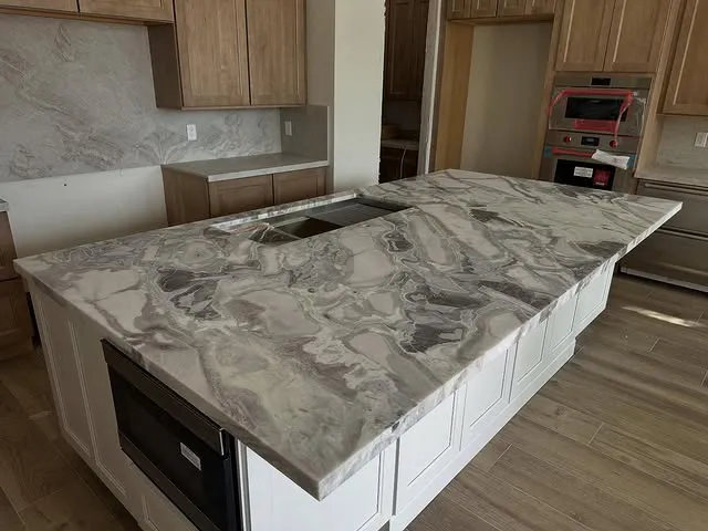 A modern kitchen featuring a large island with polished, gray and white marble countertops from Signature Stone Las Vegas, surrounded by wooden cabinets and light wood flooring.
