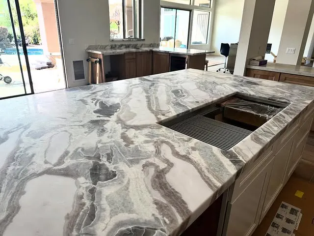 A modern kitchen with quartz countertops by Signature Stone Las Vegas, featuring a marble-patterned surface and a large sink cutout, with natural light pouring in from windows overlooking a backyard pool.