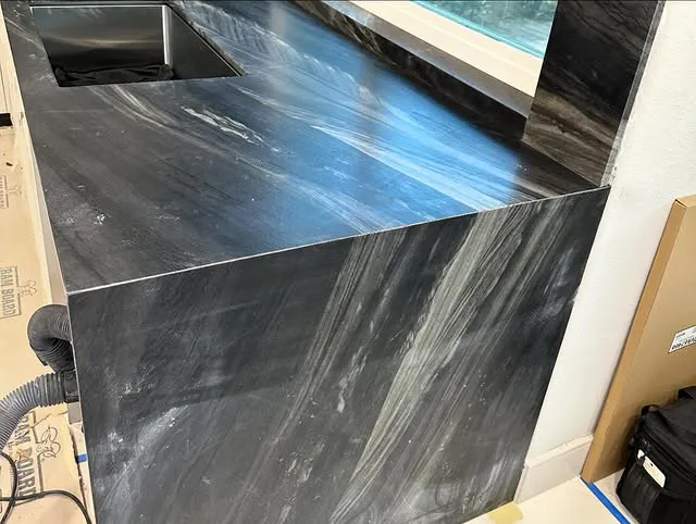 A black marble kitchen countertop with white and gray streaks, featuring an integrated sink and a matching backsplash, is shown under natural light from a nearby window—an elegant choice among modern kitchen countertops.