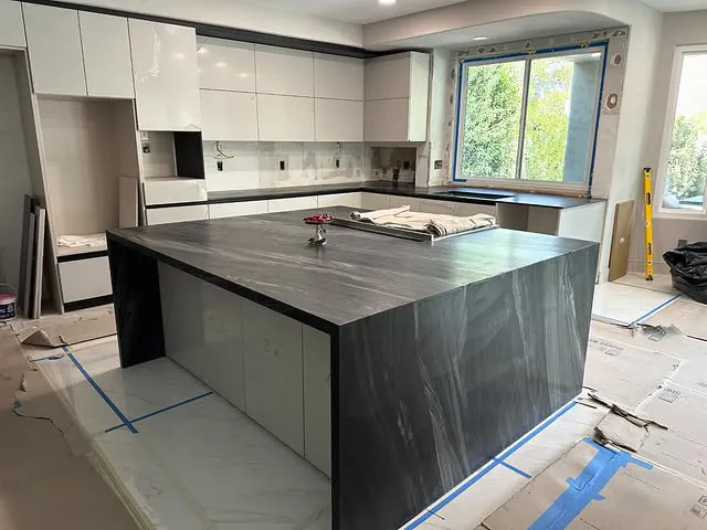 Modern kitchen under renovation with a large dark marble countertop island from Signature Stone Las Vegas, white cabinets, covered floors, and an uncovered window showing trees outside. Tools and materials are scattered around the room.
