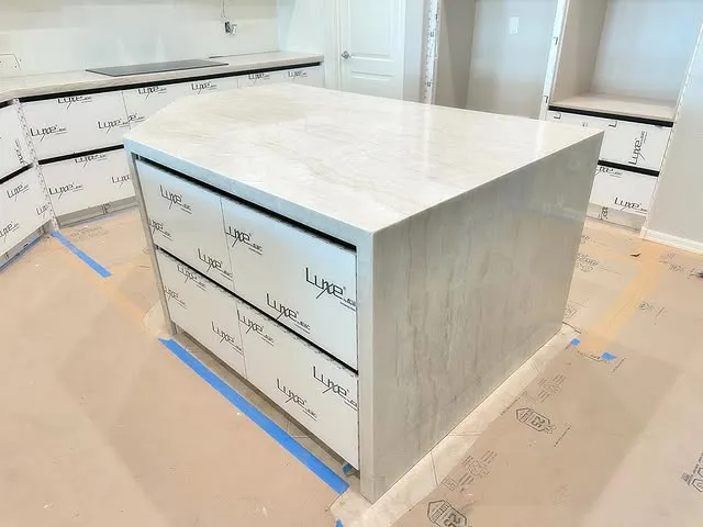 A modern kitchen island with a polished white marble countertop, perfect for showcasing premium marble countertops, features three drawers and sits in a partially finished kitchen with protective coverings and labels on the cabinets and floor.