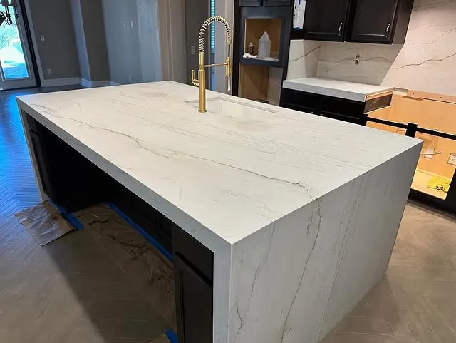 A modern kitchen island with a white marble waterfall countertop featuring subtle gray veining, a built-in sink, and a gold faucet. Dark cabinetry is visible below and around the island, highlighting the elegance of marble countertops.