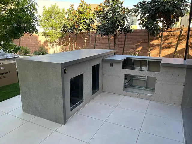 A partially completed outdoor kitchen with sleek quartz countertops and open spaces for appliances, set on white tile flooring, surrounded by trees and a brick wall in a backyard.
