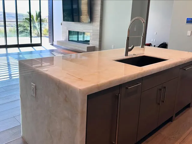 A modern kitchen island with polished marble countertops, a built-in sink with a tall curved faucet, dark cabinets, and a view of a bright open living area with large windows.