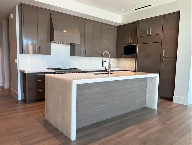 Modern kitchen with brown cabinets, stainless steel appliances, a large island with marble countertops, built-in oven, stovetop, and a sleek faucet; light wood flooring and neutral walls.