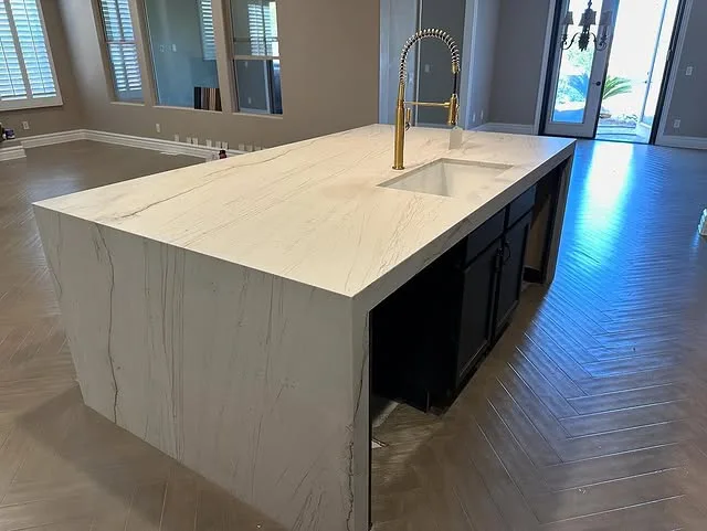 A modern kitchen island with a white marble waterfall countertop, built-in sink, tall brass faucet, and dark cabinetry sits on a light herringbone-patterned wood floor—an elegant showcase of stylish kitchen countertops in a bright, spacious room.