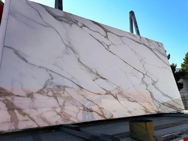 A large slab of white marble with dramatic gray and gold veining stands upright outdoors on a metal frame, perfect for stunning kitchen countertops from Signature Stone Las Vegas. A clear blue sky and some trees are visible in the background.