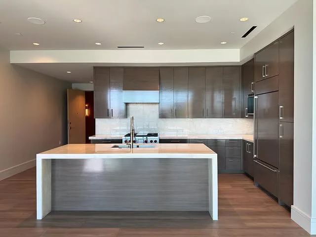 Modern kitchen with sleek brown cabinets, a large island with a sink, built-in stainless steel appliances, and light-colored quartz countertops and backsplash. The space features recessed ceiling lights and elegant wood flooring.