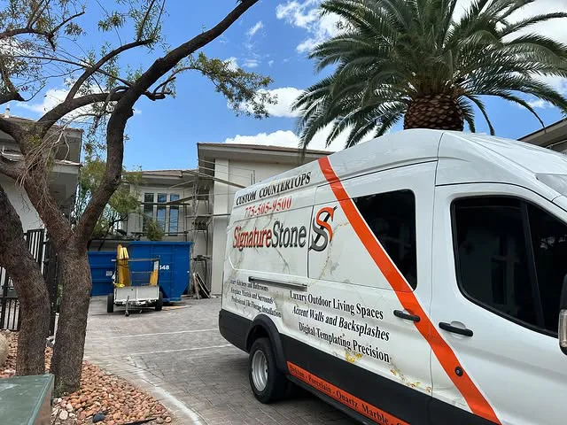A white Signature Stone van, advertising custom kitchen countertops and bathroom countertops, is parked in a driveway near a blue dumpster, with palm trees and a two-story house in the background.