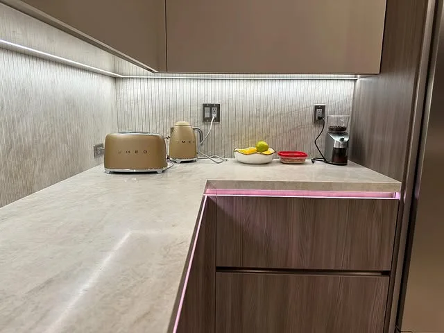 Modern kitchen countertop with quartz countertops, a beige toaster, electric kettle, bowl of fruit, red dish, and coffee maker. Under-cabinet lighting and light wood cabinets create a clean, minimalist look.